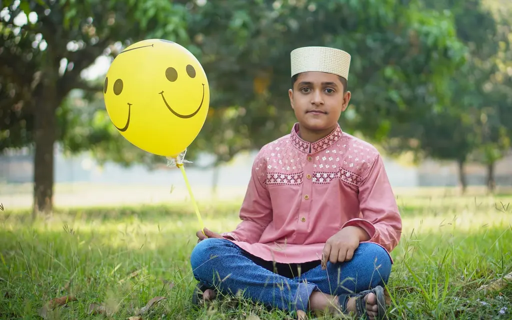 A boy in a kurta and prayer cap holding a smiley balloon